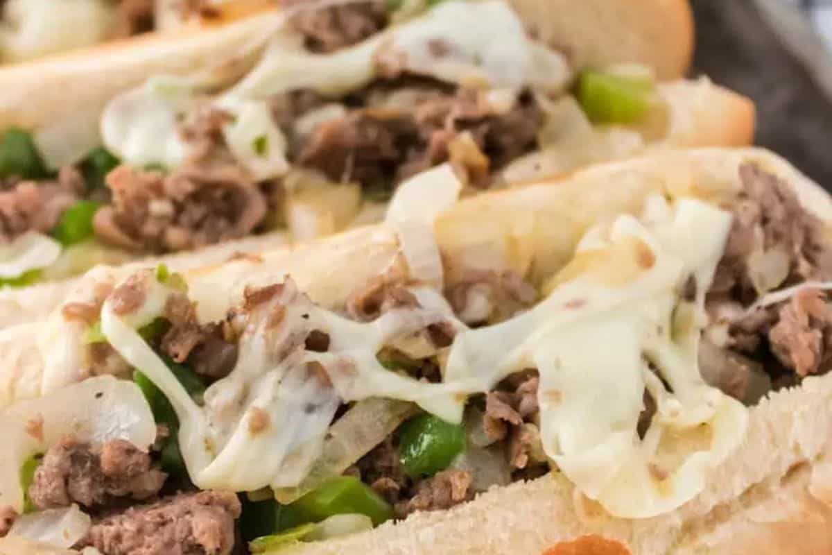 A close-up of two Philly Cheesesteak sandwiches on long rolls. The sandwiches are filled with thinly sliced, browned steak, melted white cheese, sautéed onions, and green bell peppers.