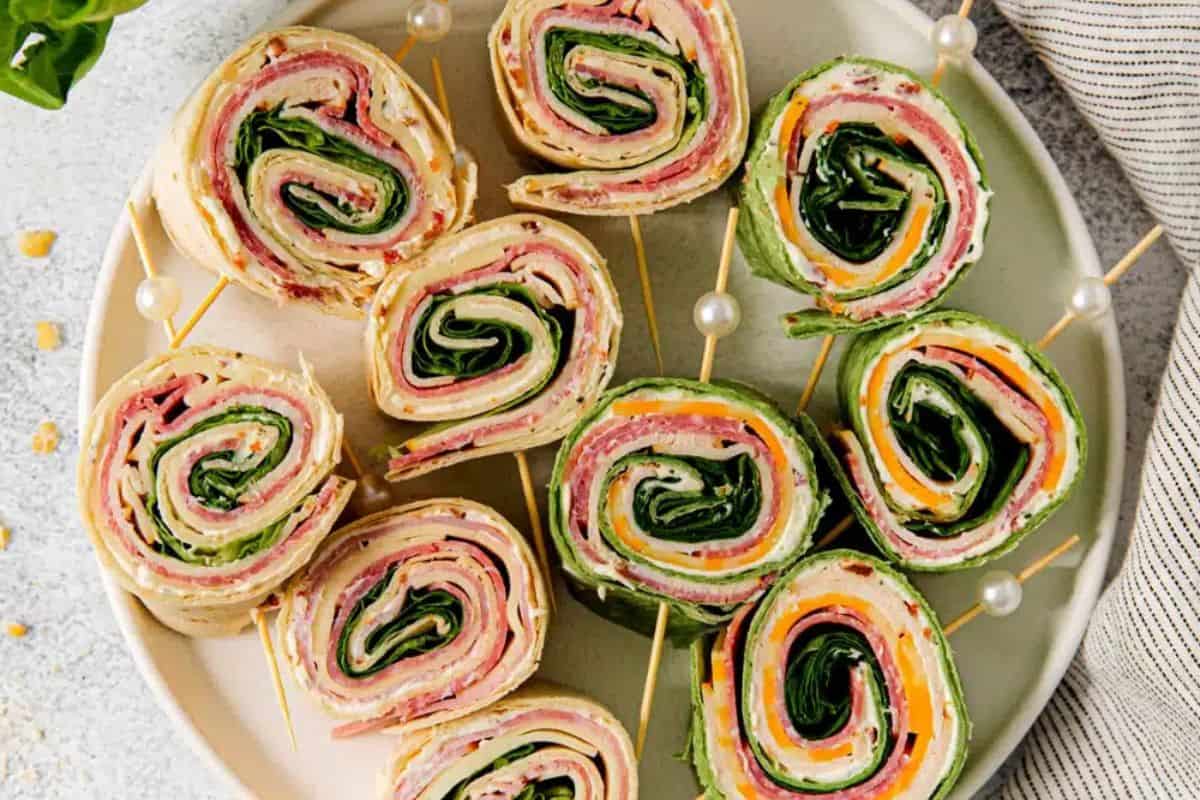 An overhead shot of a round plate filled with colorful Pinwheel Sandwiches. The spirals show layers of tortillas, creamy spread, ham and salami, cheese, and green leafy vegetables. 