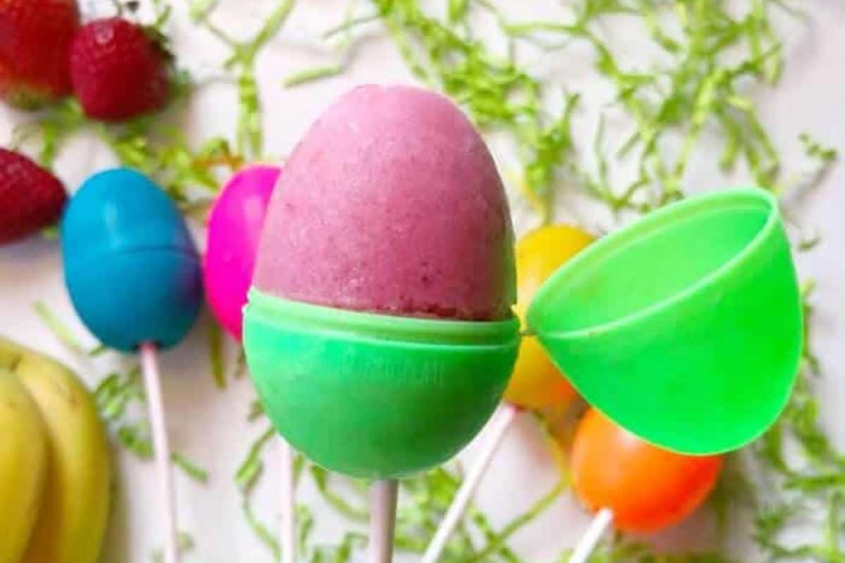 A pink popsicle shaped like an egg is held in a green plastic egg, with other colorful eggs and fruit in the background—perfect for Easter egg ideas or easy Easter crafts.