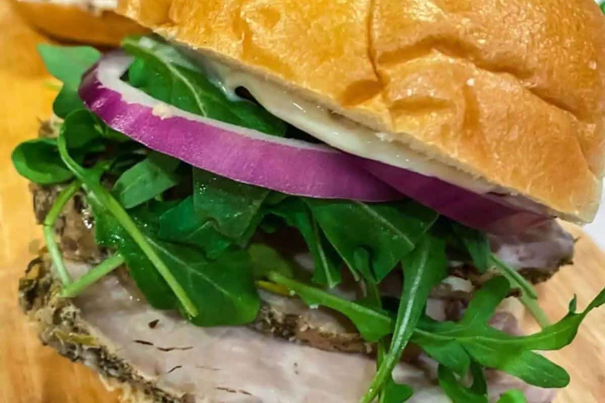  close-up of a Porchetta Sandwich on a wooden surface. Slices of seasoned and roasted pork are layered in a soft roll with fresh arugula leaves and thin slices of red onion. A creamy sauce is visible on the top bun.