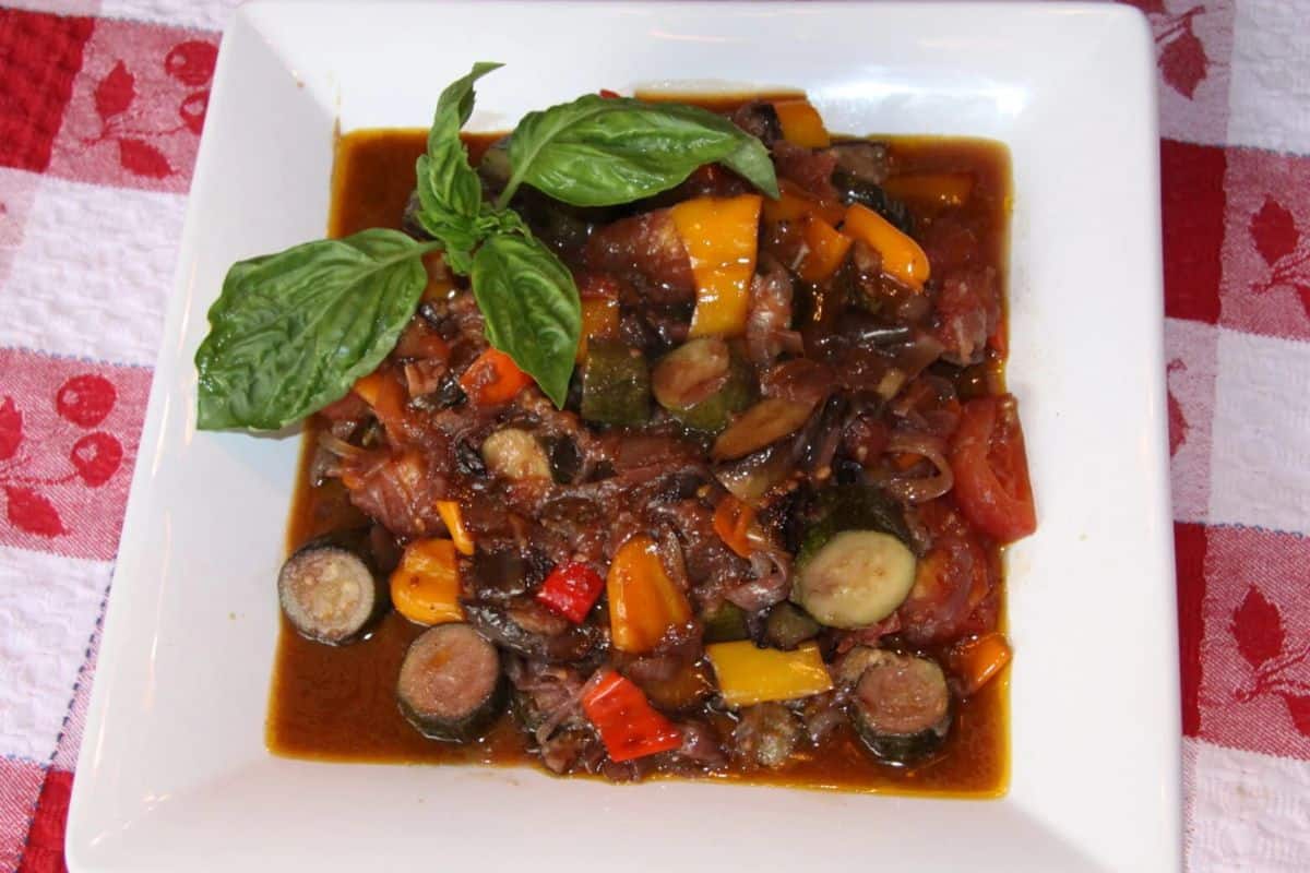 An overhead shot of a square white plate filled with Ratatouille. The stew features colorful chunks of eggplant, zucchini, red, yellow, and orange bell peppers, onions, and tomatoes in a rich sauce, garnished with fresh basil leaves.