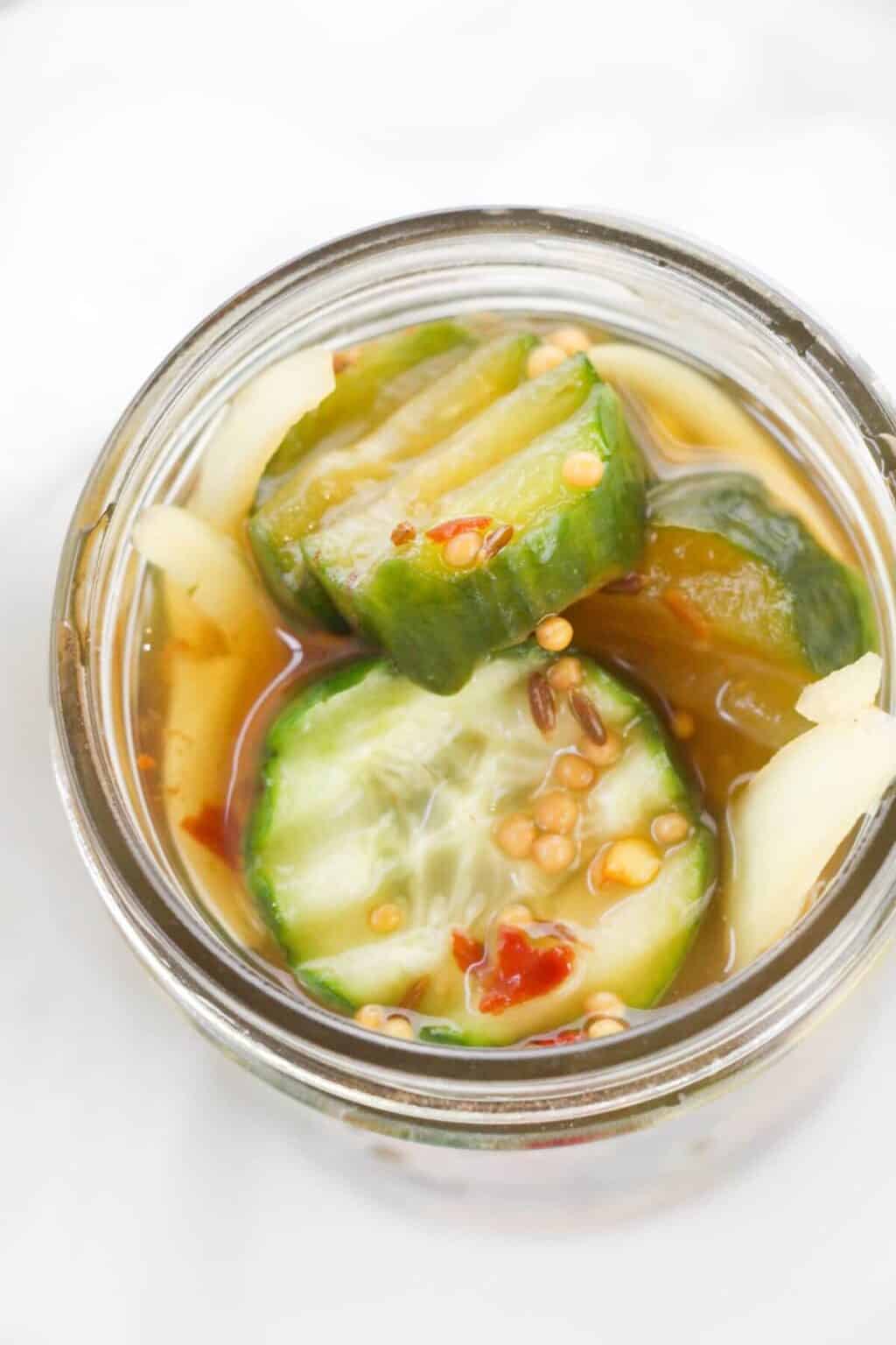 A jar of Refrigerator Bread and Butter Pickles in brine, featuring sliced pickles with visible mustard seeds, spices, and onion slices.