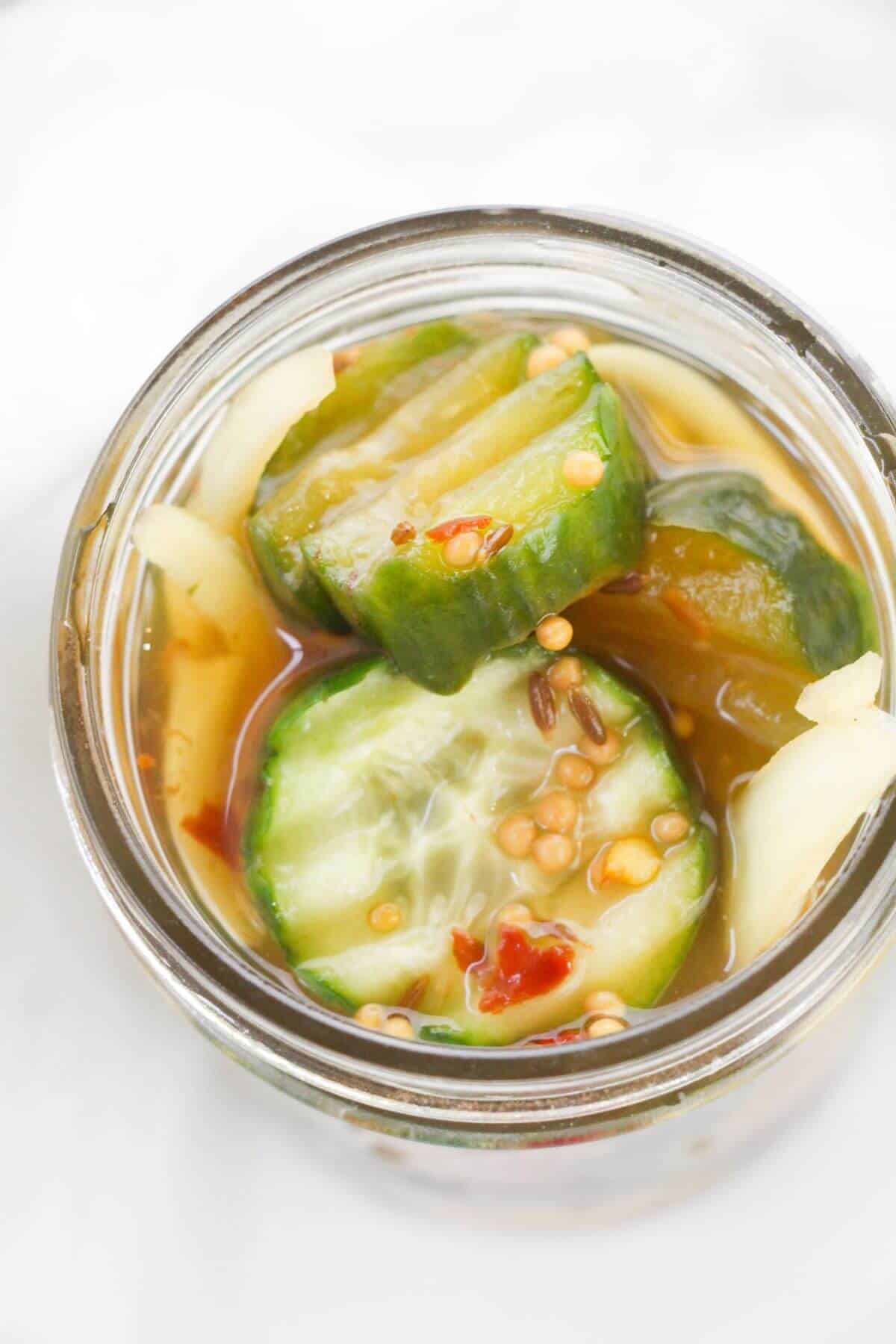 A jar of Refrigerator Bread and Butter Pickles in brine, featuring sliced pickles with visible mustard seeds, spices, and onion slices.