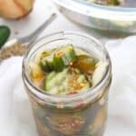 A glass jar cucumbers and onions in brine—sits on a white surface, with a bowl and an onion in the background.
