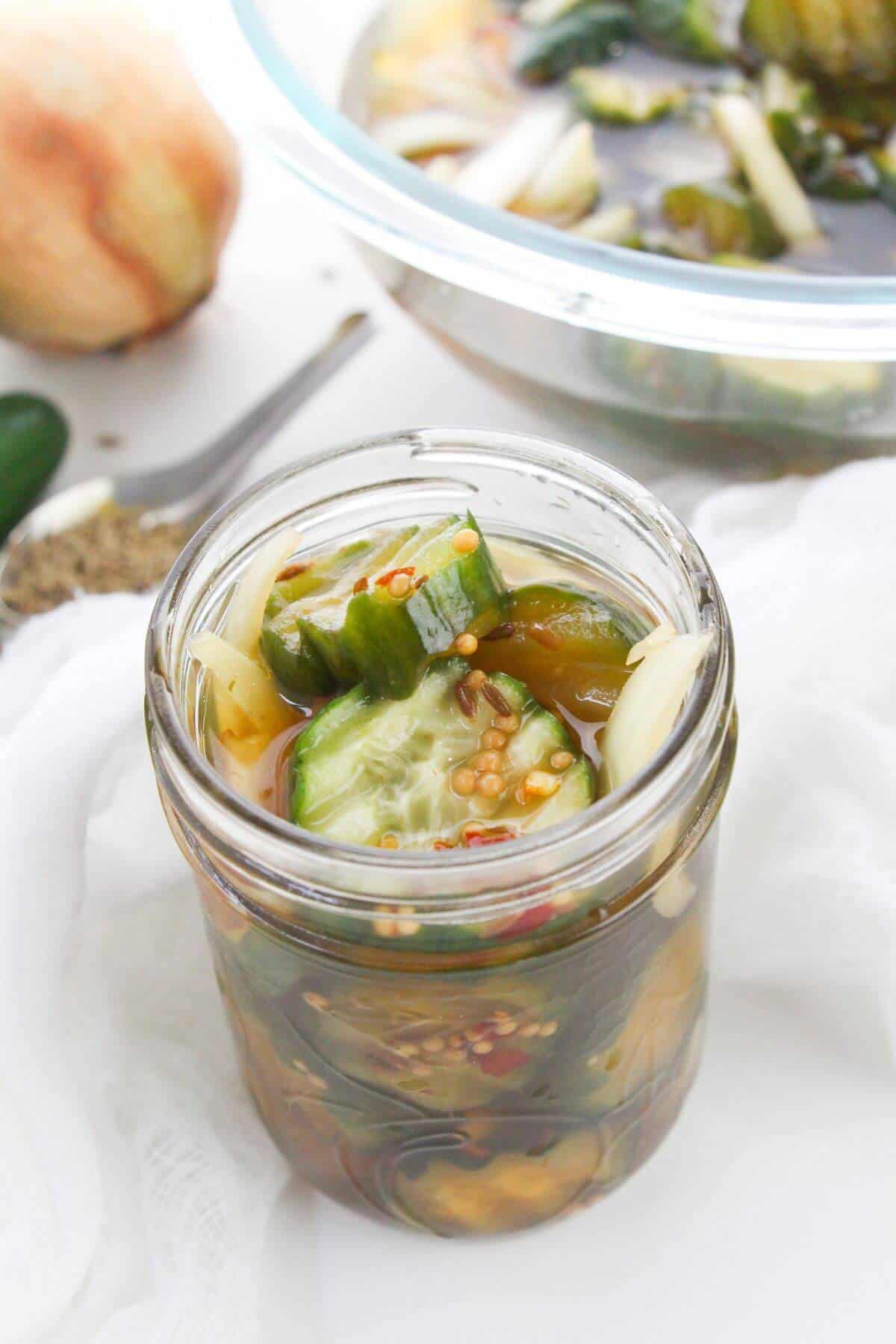 A glass jar cucumbers and onions in brine—sits on a white surface, with a bowl and an onion in the background.