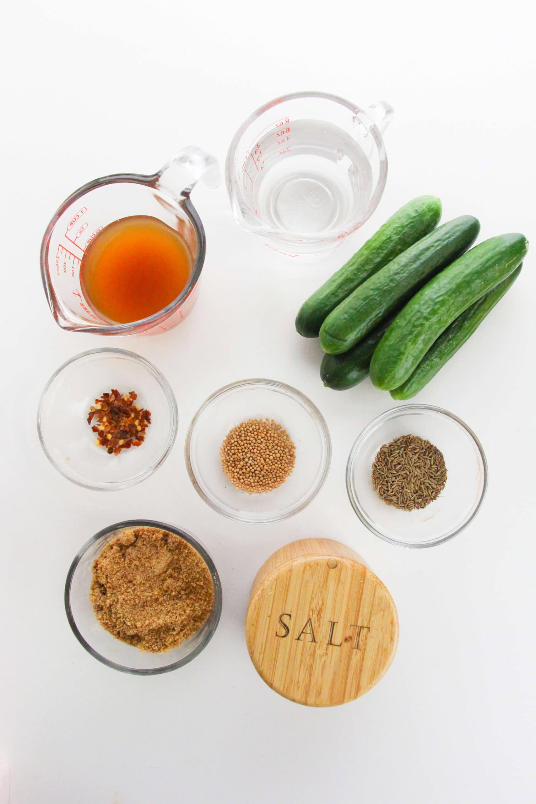 Ingredients on a white surface: measuring cups with vinegar and water, four cucumbers, small bowls of red pepper flakes, mustard seeds, cumin seeds, brown sugar, and a salt container.