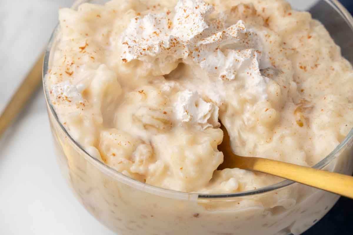 A bowl of creamy rice pudding, a classic cuisine favorite, topped with whipped cream and a sprinkle of cinnamon, with a gold spoon resting in the bowl.