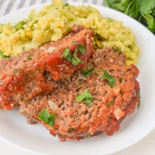 A plate featuring two slices of meatloaf topped with ketchup and parsley, served alongside creamy mashed potatoes mixed with herbs, delivers a classic comfort food experience. The recipe calls for a dash of creativity: try crushed Ritz Crackers for extra texture.