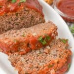 Sliced meatl on a white platter, topped with tomato sauce and herbs. Accompanied by a bowl of ketchup.