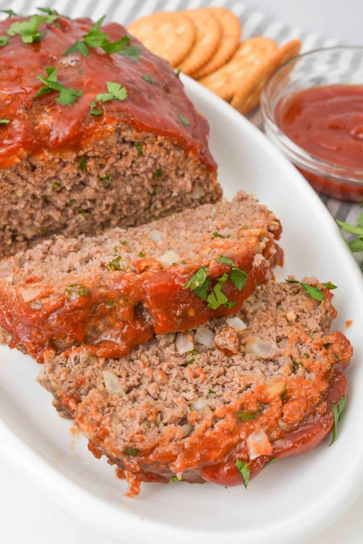 Sliced meatl on a white platter, topped with tomato sauce and herbs. Accompanied by a bowl of ketchup.