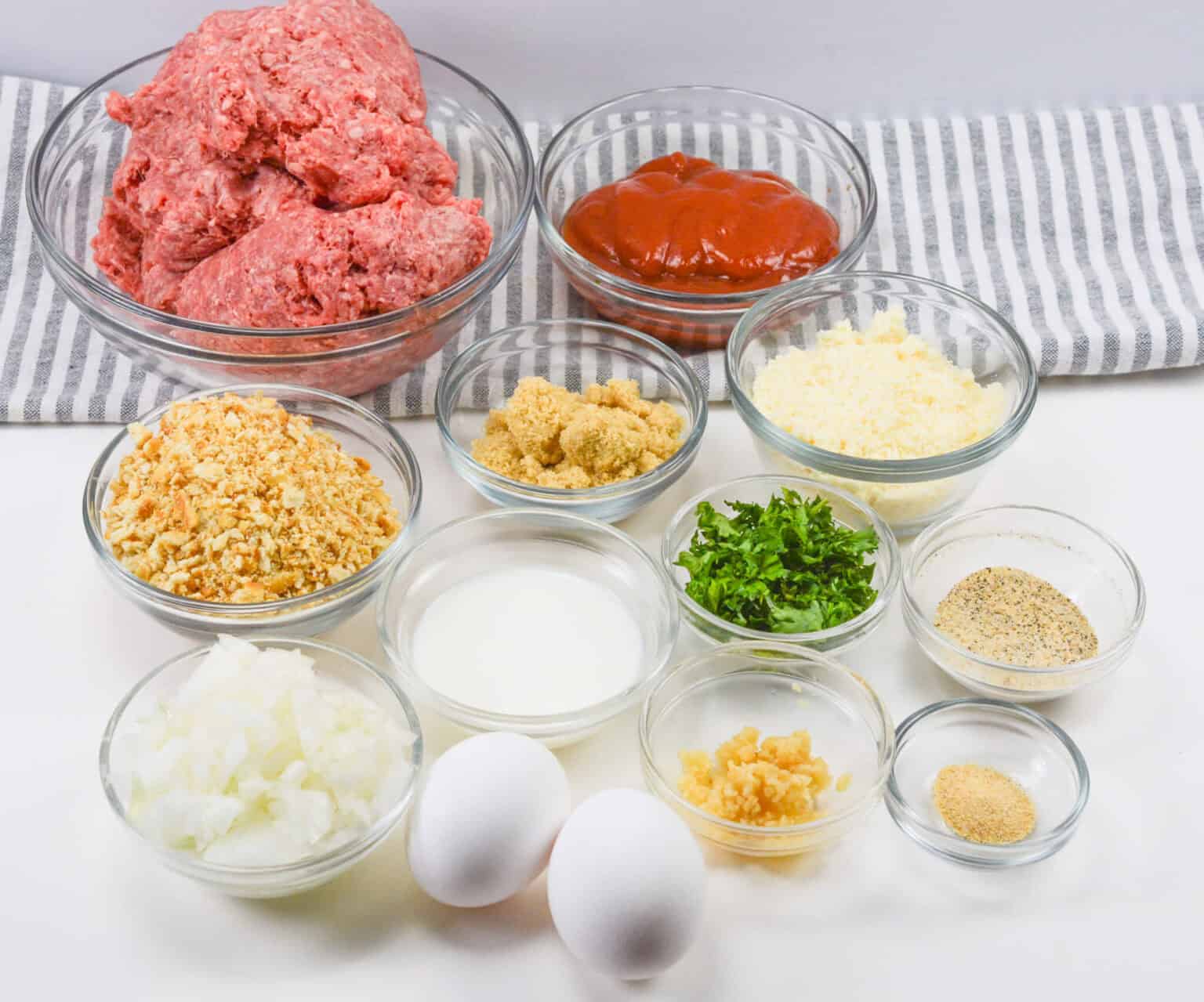 Ingredients for a Ritz Cracker meatloaf recipe in glass bowls: ground beef, ketchup, crushed Ritz Crackers, brown sugar, grated cheese, chopped parsley, minced onion, milk, eggs, garlic, and seasonings.