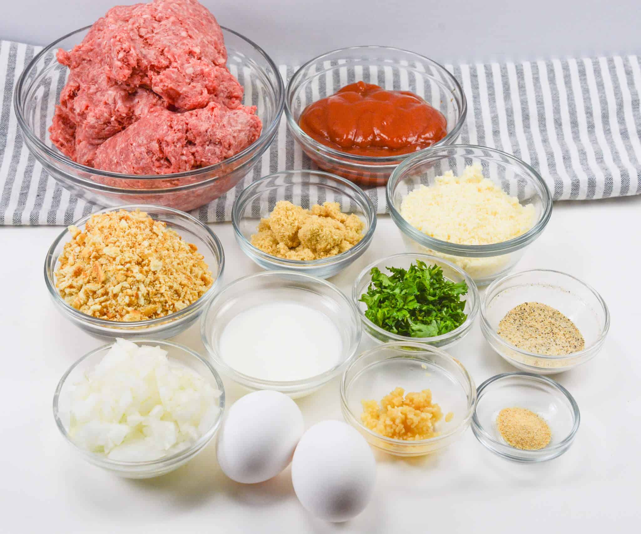Ingredients for a Ritz Cracker meatloaf recipe in glass bowls: ground beef, ketchup, crushed Ritz Crackers, brown sugar, grated cheese, chopped parsley, minced onion, milk, eggs, garlic, and seasonings.