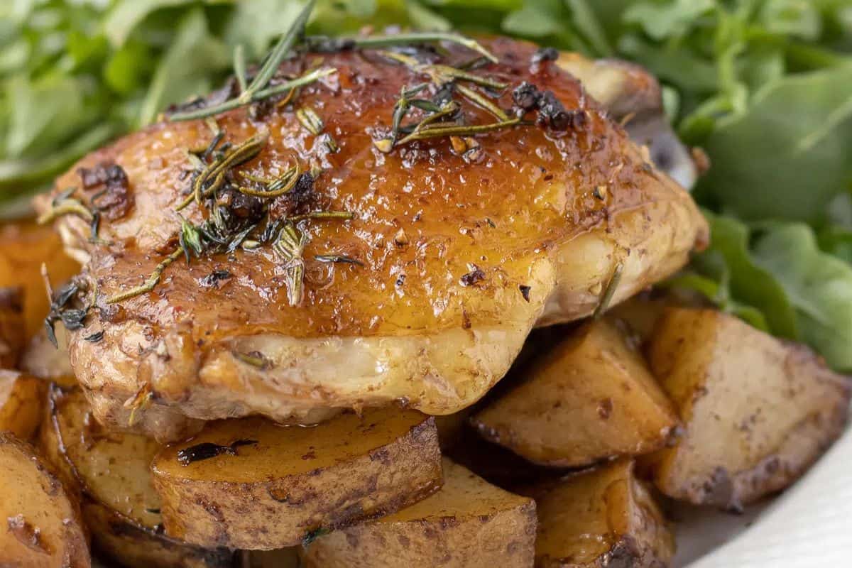 Roasted juicy chicken thighs with herbs on seasoned potato wedges, served alongside fresh leafy greens—perfect for fans of hearty chicken thigh recipes.