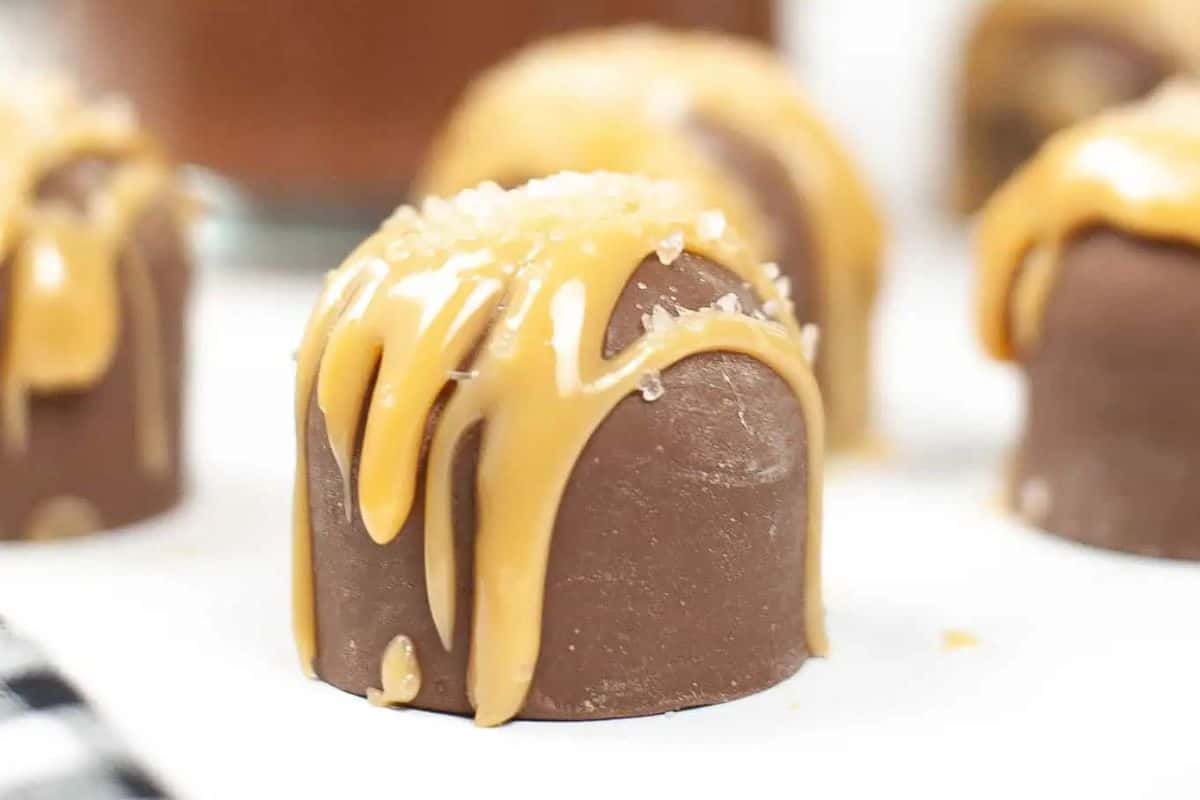 A close-up of a round hot chocolate bomb drizzled with caramel sauce and sprinkled with coarse salt, showcasing indulgent desserts, with more candies blurred in the background.