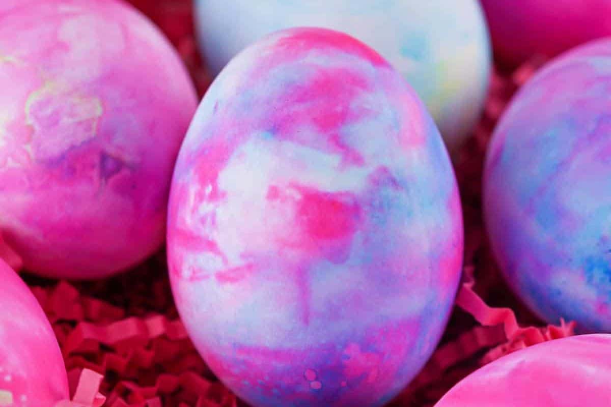 A close-up of vibrantly colored Easter eggs with pink, blue, and purple marbled patterns showcases creative Easter egg decorating, with the eggs nestled on a bed of pink shredded paper.