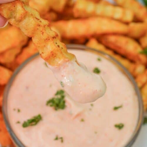 A crinkle-cut French fry is dipped in creamy Sheetz Boom Boom Sauce with herbs, with more fries and sauce visible on a white plate in the background.