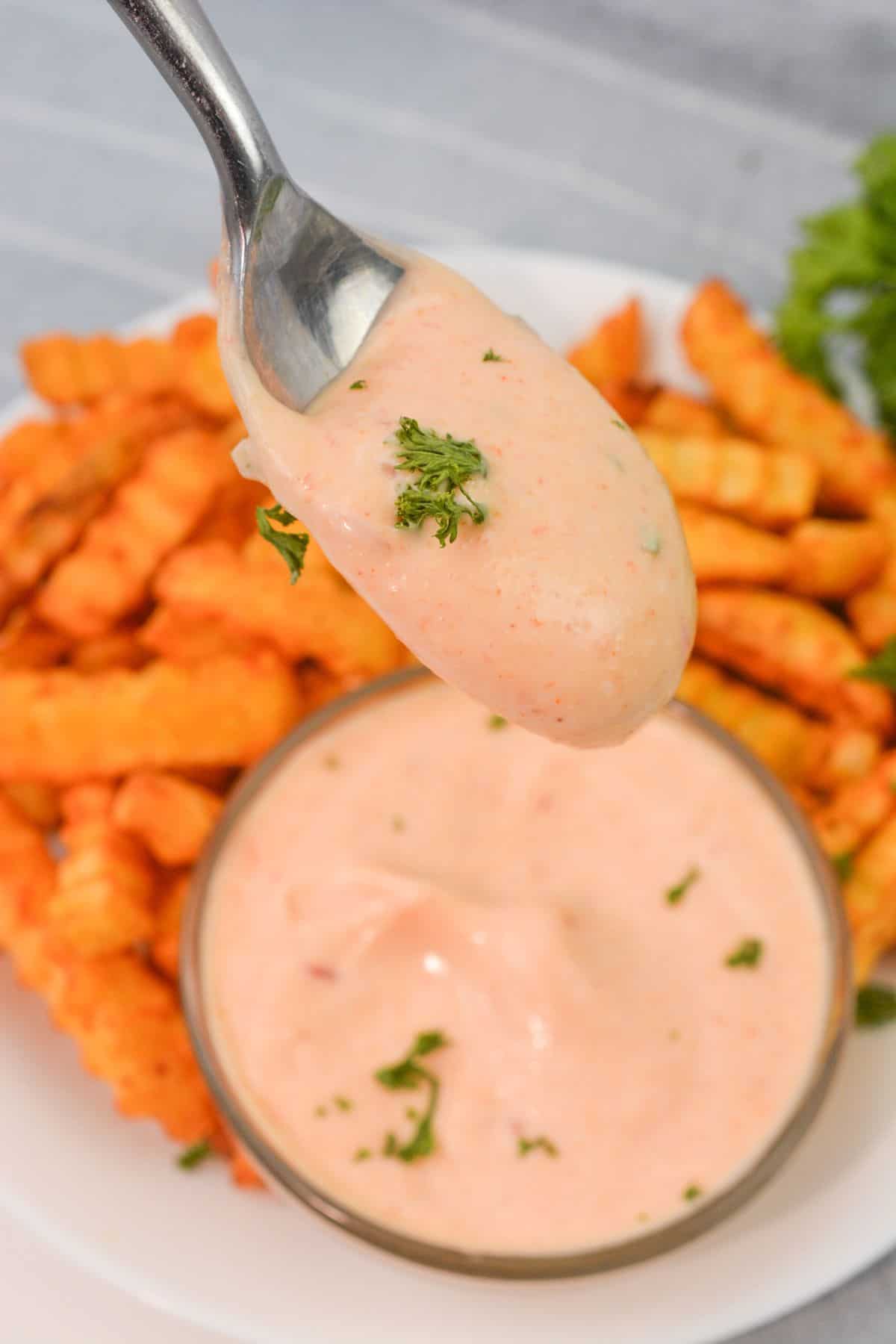 A spoon holds Sheetz Boom Boom Sauce, the pink dipping sauce flecked with green herbs, above a bowl of the same sauce, with crinkle-cut fries on a plate in the background.