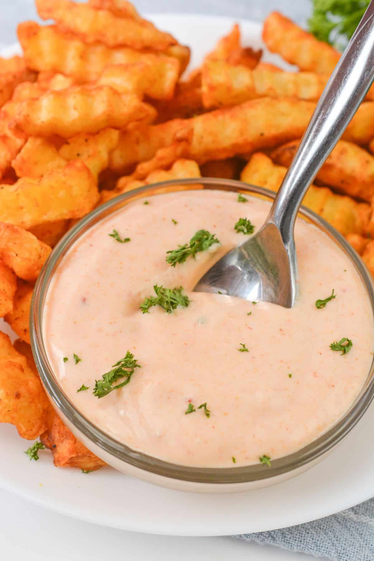 A bowl of creamy Sheetz Boom Boom Sauce dipping sauce, garnished with chopped herbs, sits on a plate beside crinkle-cut fries, with a spoon in the sauce.