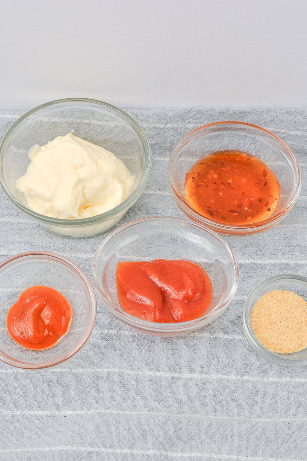Five glass bowls containing mayonnaise, a red sauce, ketchup, Boom Boom Sauce from Sheetz, and a yellow powder are arranged on a striped cloth.