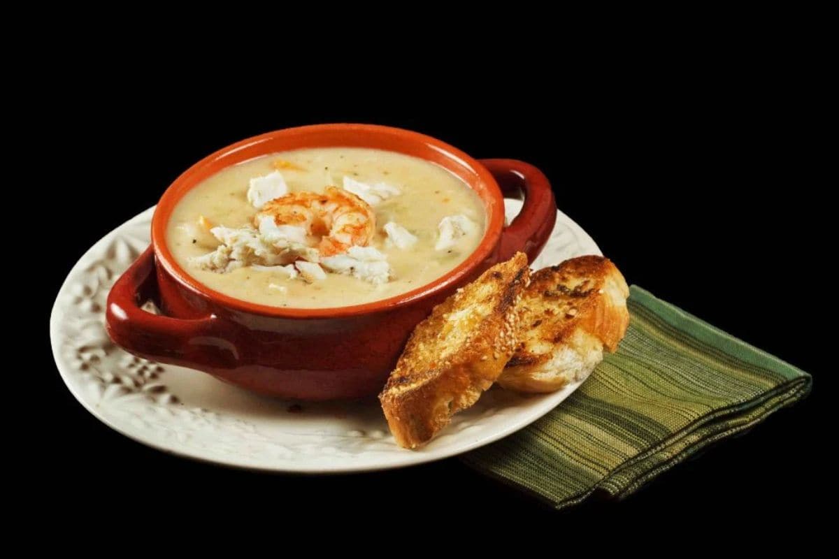 A bowl of creamy soup garnished with crab meat and shrimp, served with two slices of toasted bread on a white plate and a green napkin—an ode to classic meals that never go out of style.