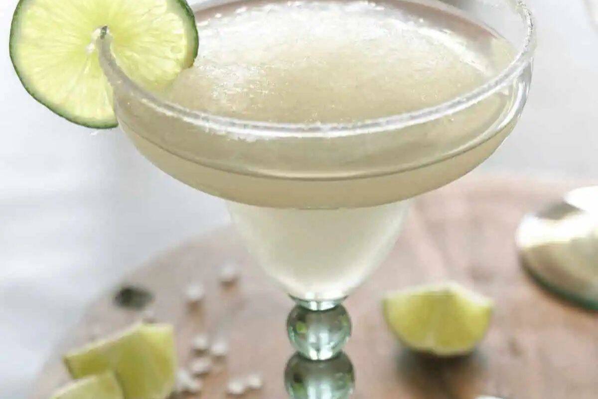 A margarita in a stemmed glass with a salted rim, garnished with a lime slice, sits on a wooden surface with lime wedges nearby—a true classic cocktail.