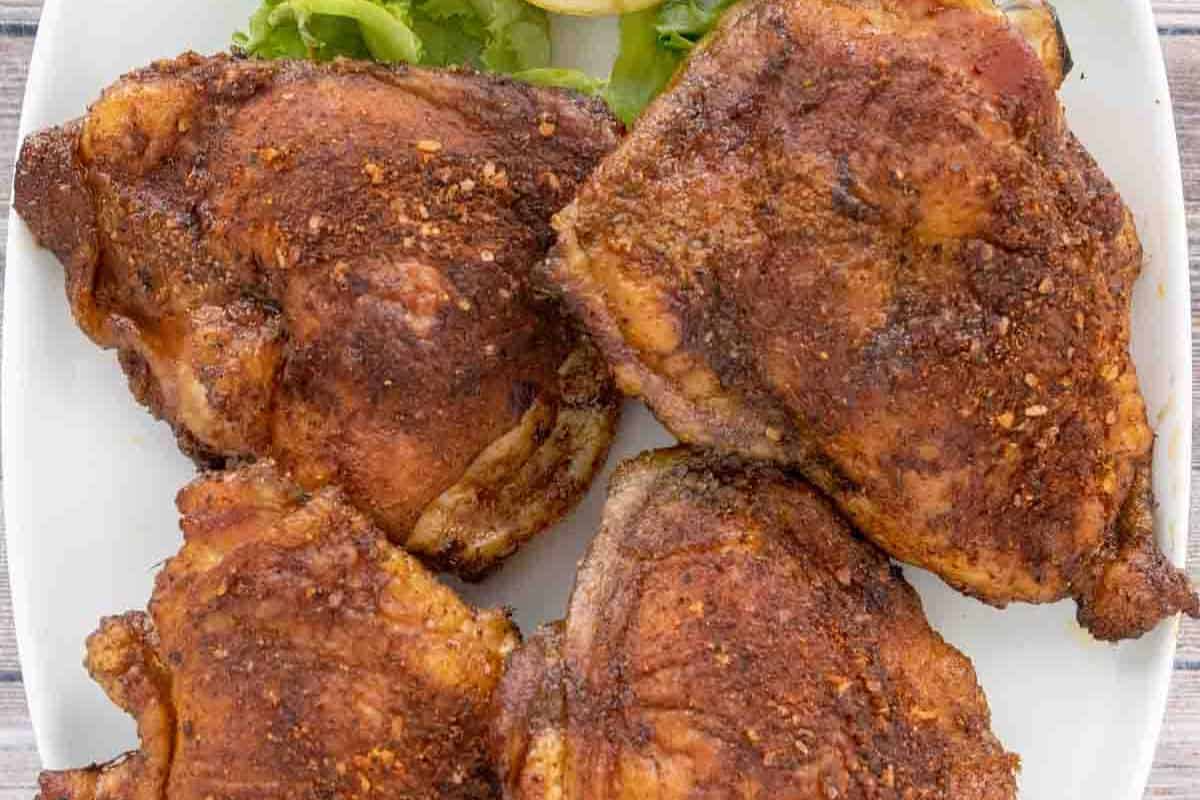 Four seasoned, cooked juicy chicken thighs are arranged on a white plate with a small garnish of lettuce and onion in the background—perfect inspiration for your favorite chicken thigh recipes.