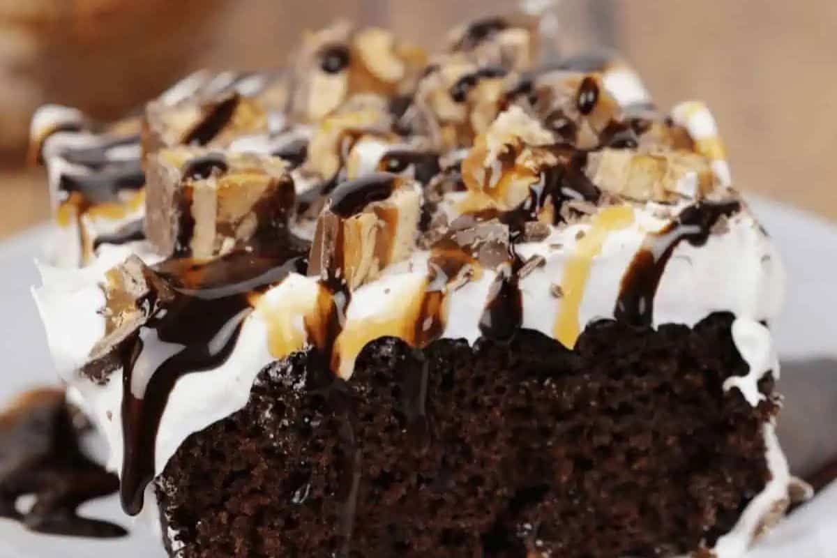 A slice of Snickers poke cake topped with whipped cream, chopped candy bars, and drizzled with chocolate and caramel sauce—perfect for fans of indulgent desserts and those looking for easy caramel recipes.