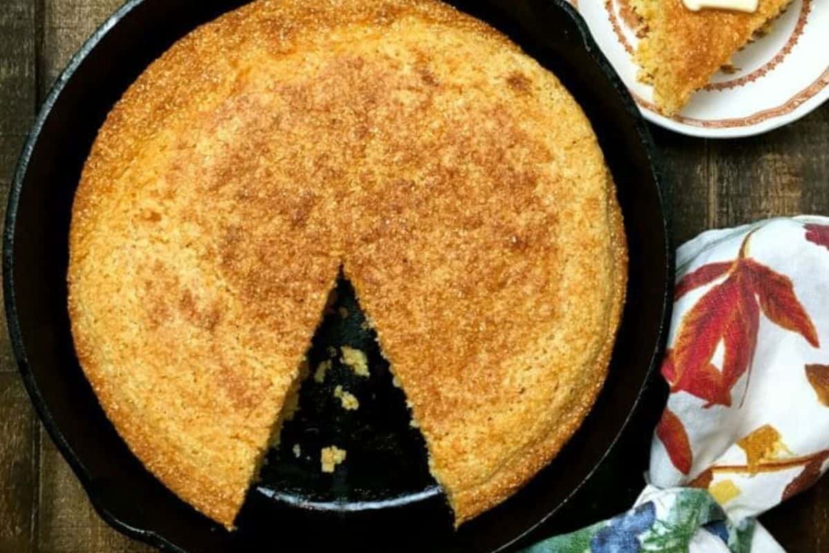 A round cornbread with a slice removed sits in a black cast iron skillet, next to a plate holding the cut slice, on a wooden surface—an inviting display of classic recipes and timeless meals.