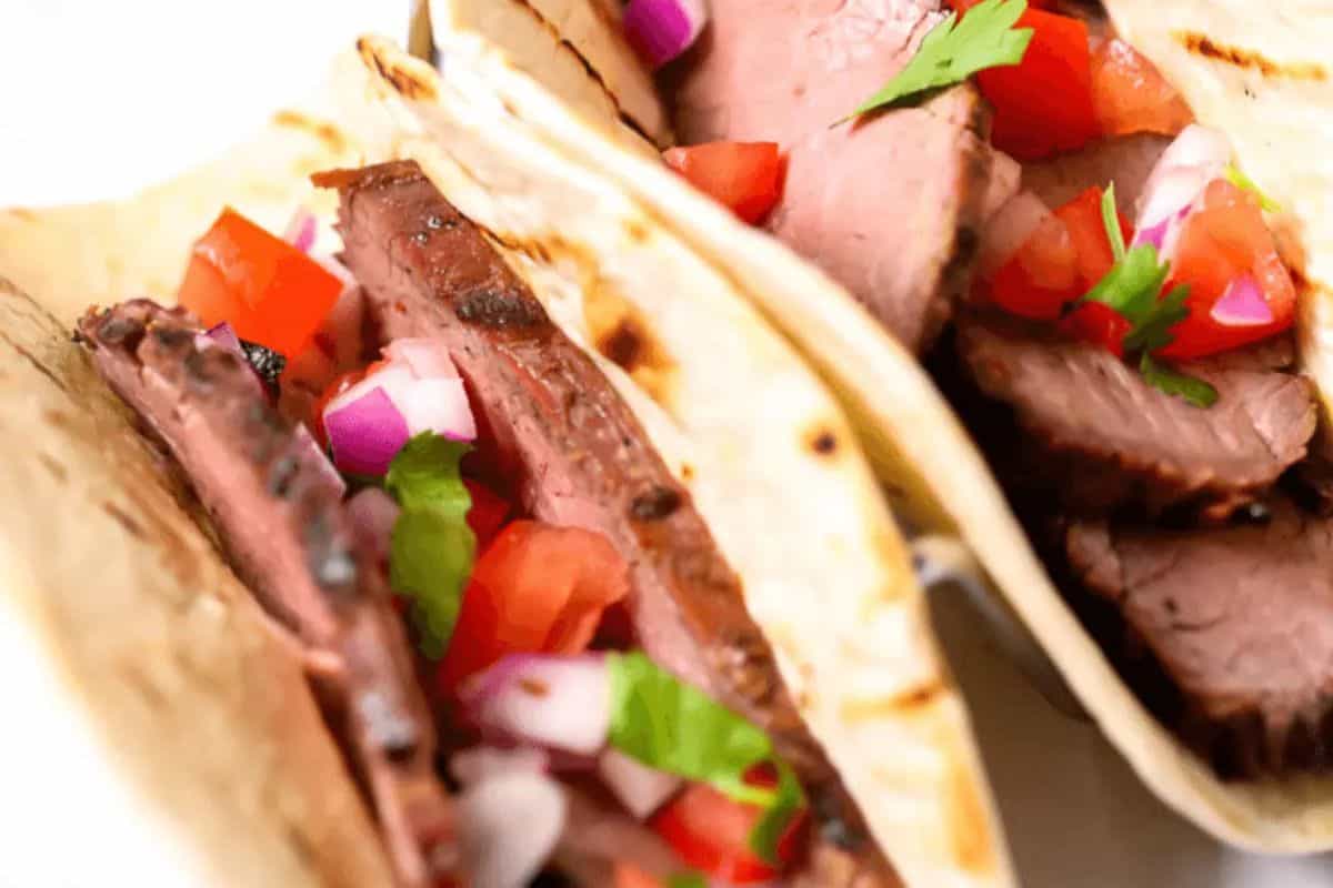 Two tacos filled with slices of grilled meat, diced tomatoes, red onions, and cilantro, wrapped in soft tortillas—one of those delicious meals perfect for dinner ideas that are simple yet flavorful.