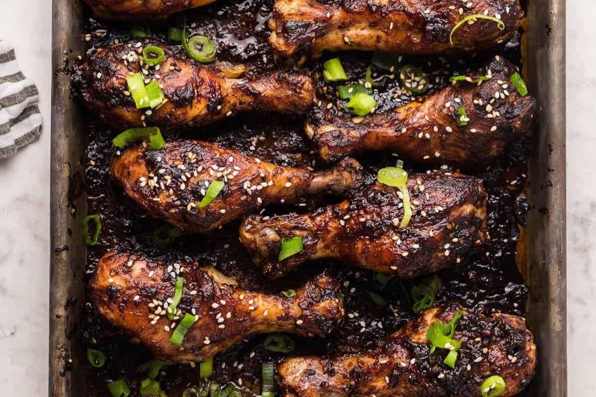 Baked chicken drumsticks coated in a dark glaze, garnished with sesame seeds and sliced green onions on a baking tray—perfect for anyone craving finger licking chicken or searching for new chicken drumstick recipes.