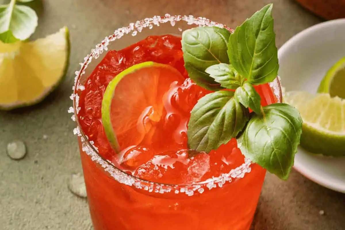 A close-up shot of a vibrant red Strawberry Basil Margarita in a glass with a salt-rimmed edge. The drink is garnished with a lime slice and fresh basil leaves.