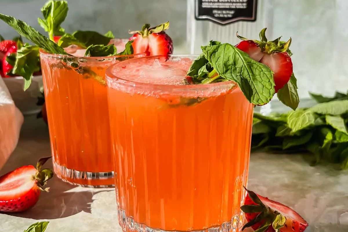 Two glasses of a bright orange-red drink, inspired by classic cocktails, are garnished with fresh mint and strawberries on a table, with extra strawberries and mint leaves nearby.
