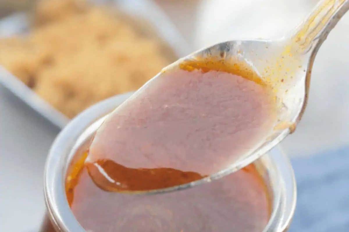 A close-up shot of a spoonful of Texas BBQ Sauce, a thick, reddish-brown glaze, being lifted from a glass jar.