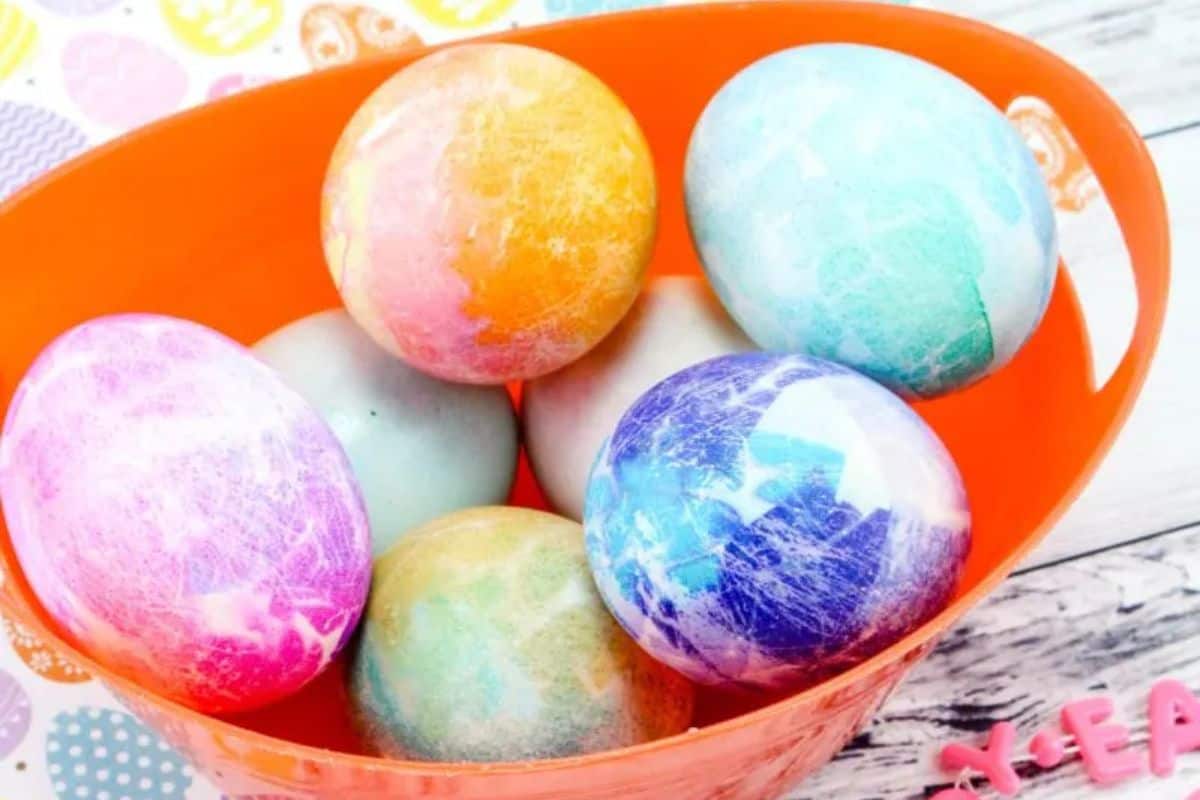 An orange bowl holds seven brightly colored, marbled Easter eggs on a white wooden surface with pastel decorations nearby—perfect inspiration for Easter Egg Decorating and Easy Easter Crafts.