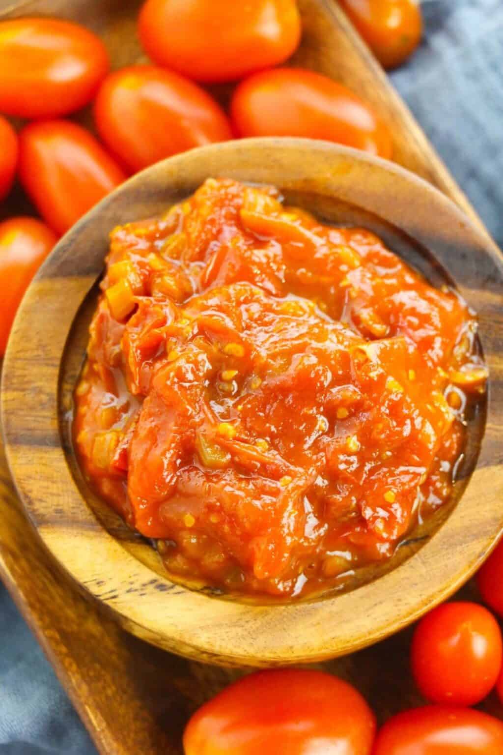 A wooden bowl filled with chunky tomato sauce, reminiscent of savory tomato jam, surrounded by whole grape tomatoes on a wooden tray.
