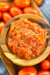A wooden bowl filled with chunky tomato sauce, reminiscent of savory tomato jam, surrounded by whole grape tomatoes on a wooden tray.