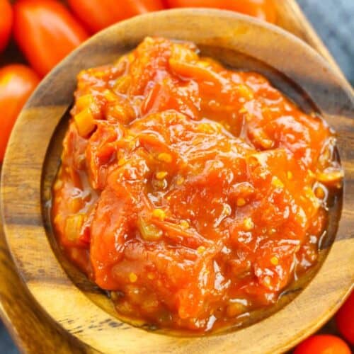 A wooden bowl filled with chunky tomato sauce, reminiscent of savory tomato jam, surrounded by whole grape tomatoes on a wooden tray.