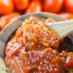 A wooden spoon scoops the condiment from a wooden bowl, with whole tomatoes blurred in the background.