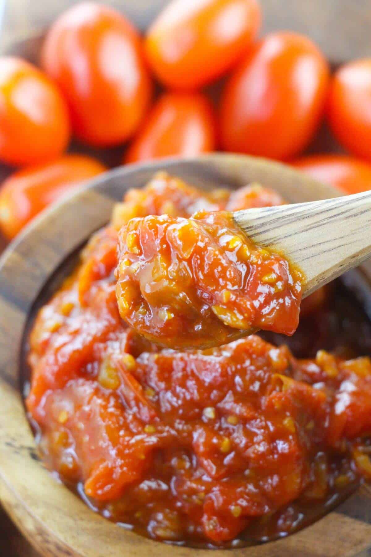A wooden spoon scoops the condiment from a wooden bowl, with whole tomatoes blurred in the background.