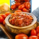 A wooden bowl filled with chunky tomato relish, reminiscent of tomato jam, sits on a tray surrounded by fresh grape tomatoes, with a blue basket and an onion in the background.