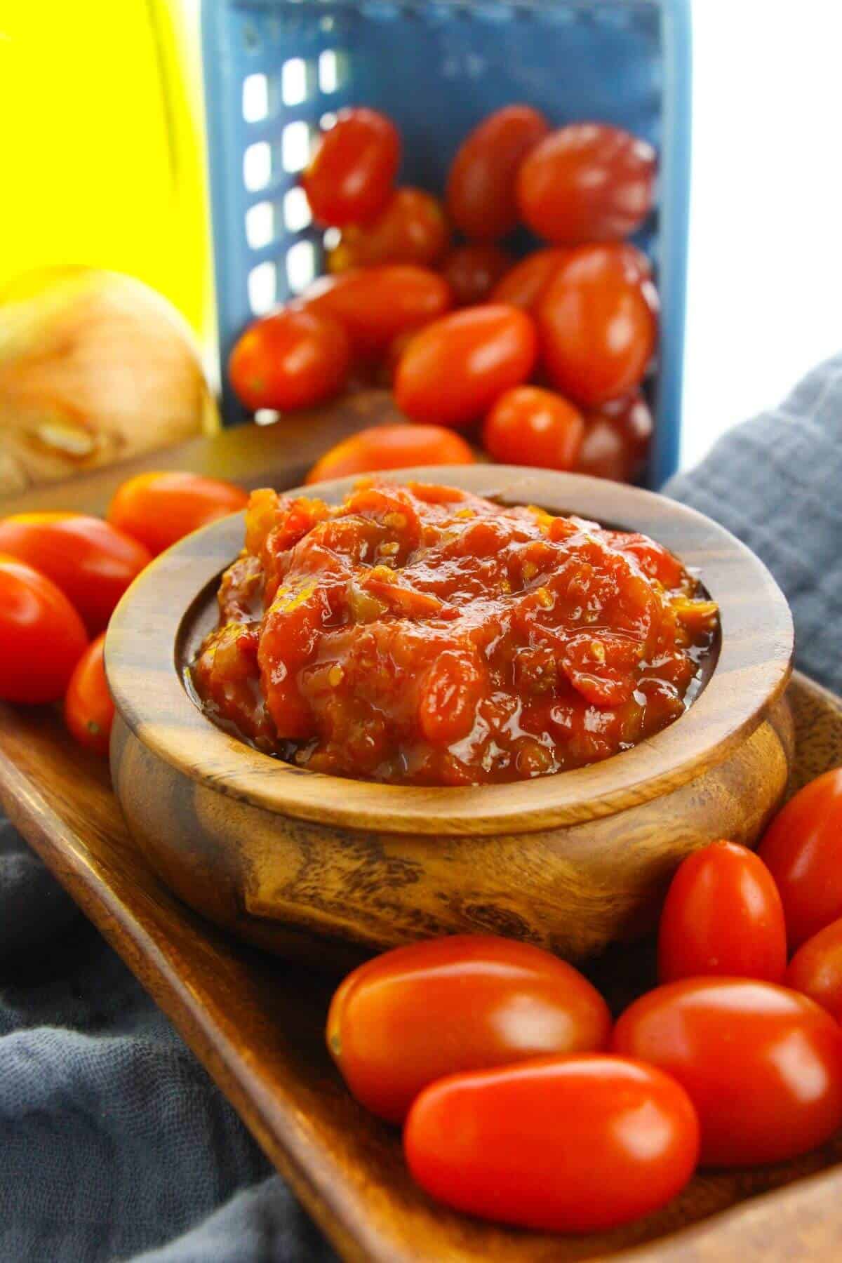 A wooden bowl filled with chunky tomato relish, reminiscent of tomato jam, sits on a tray surrounded by fresh grape tomatoes, with a blue basket and an onion in the background.