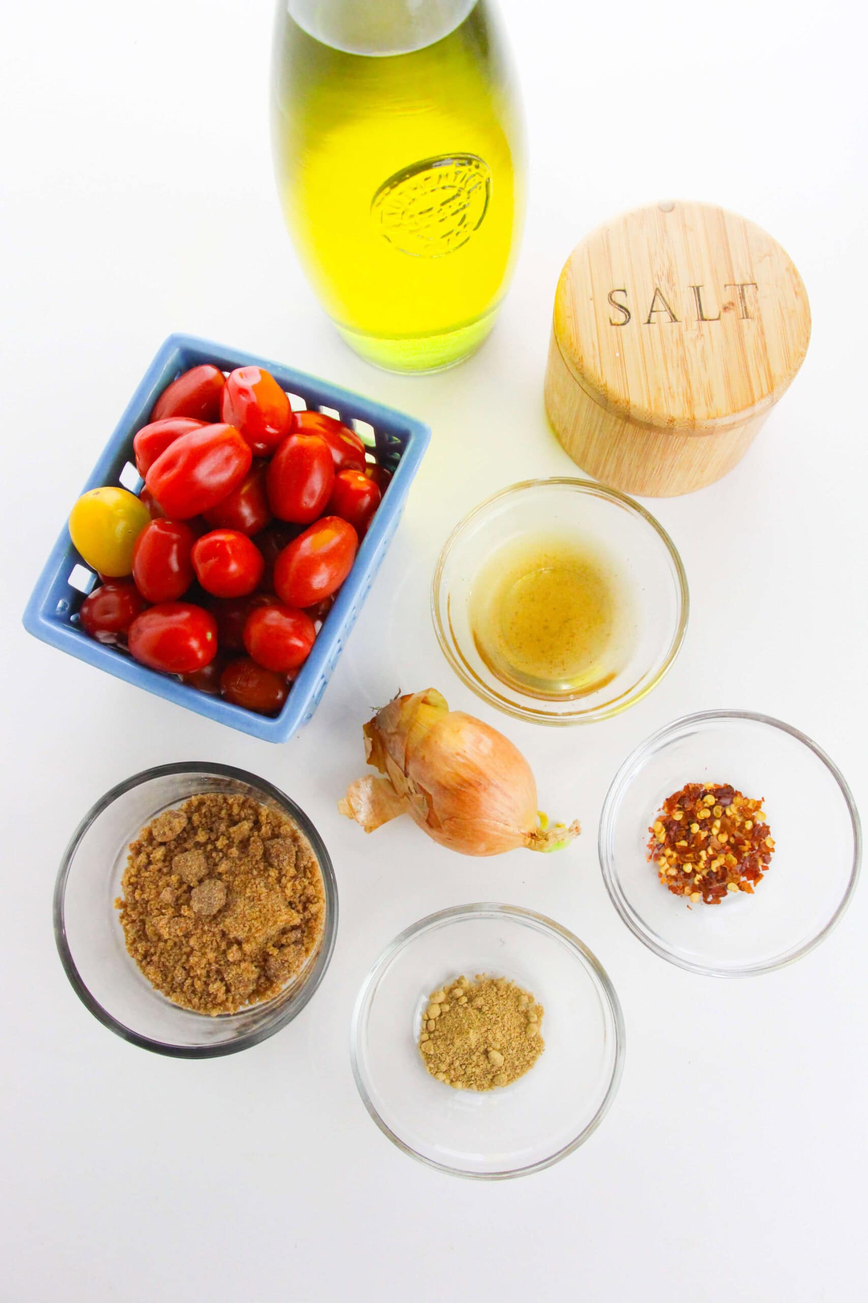 Overhead view of ingredients for Tomato Jam—olive oil, salt, cherry tomatoes, shallot, vinegar, brown sugar, ground spice, and red pepper flakes—all beautifully arranged in bowls on a white surface.