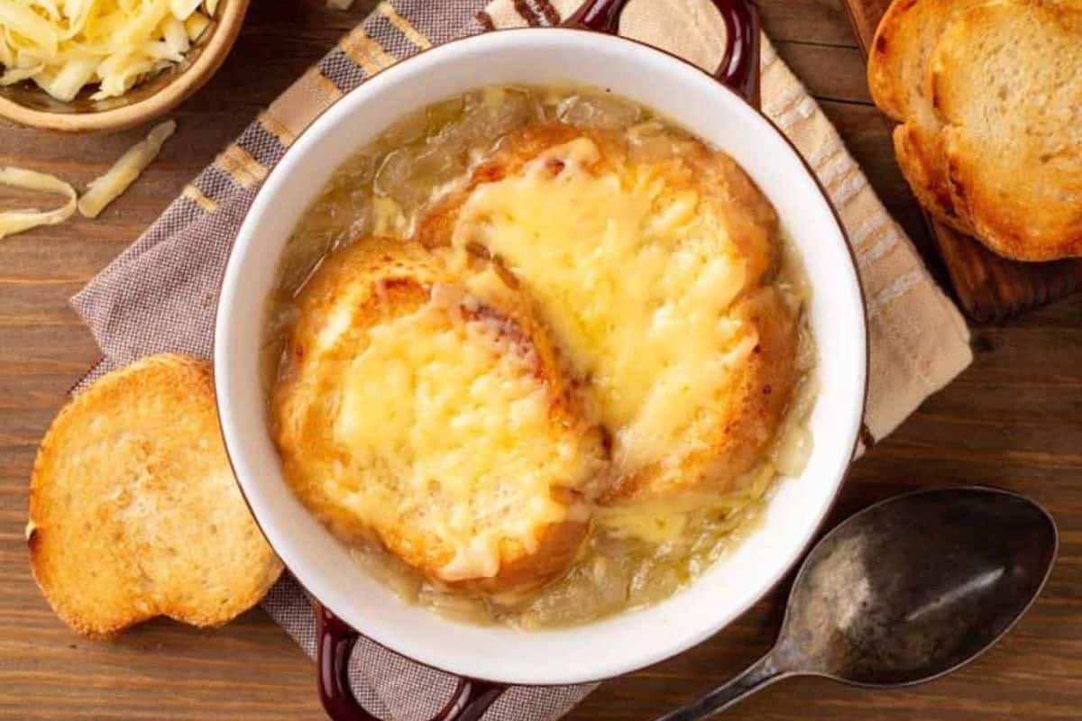 An overhead shot of a white bowl of Traditional French Onion Soup. Caramelized onions are visible in a clear broth, topped with two slices of toasted bread covered in melted, golden-brown cheese.