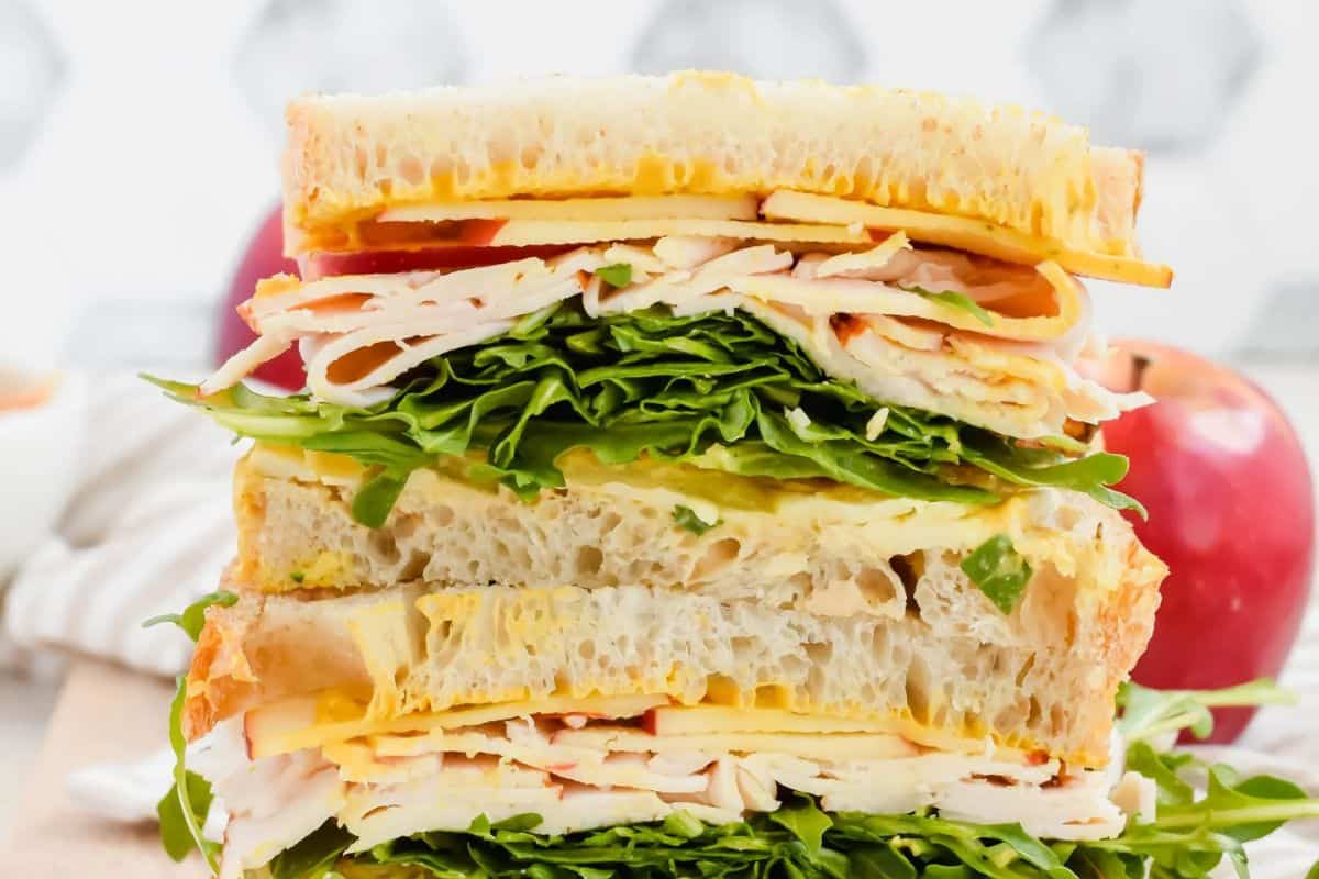  A close-up of a thick Turkey Apple Cheddar Sandwich, cut in half to show the layers of sliced turkey, white cheddar cheese, thin slices of red apple, and fresh green arugula between two slices of bread.