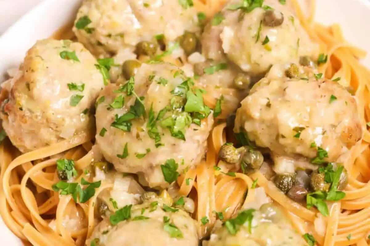 A close-up overhead shot of a white bowl filled with Veal Piccata Meatballs served over pasta. The meatballs are coated in a creamy, light-colored sauce with capers and garnished with fresh parsley.