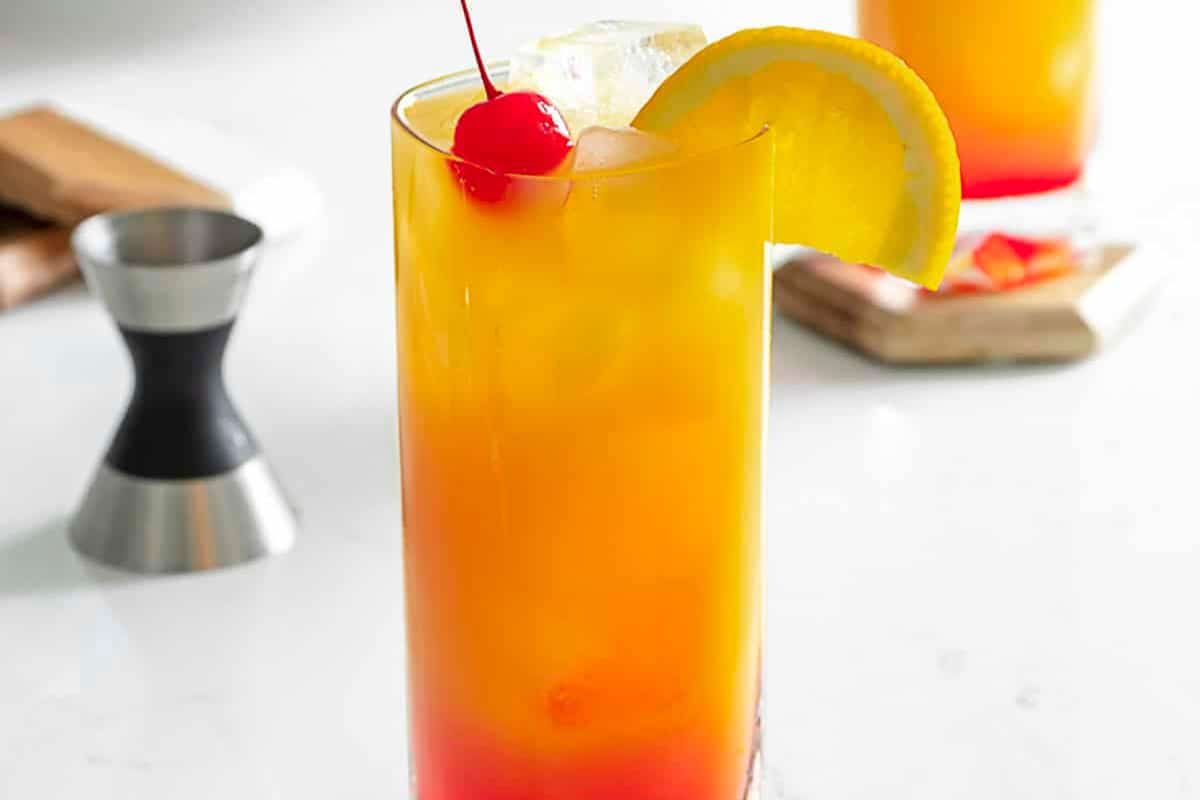 A tall glass of orange and red layered drink with ice, garnished with a cherry and orange slice, sits on a white surface—one of those timeless cocktails everyone should know.