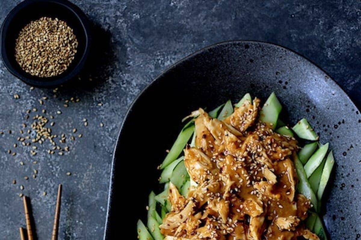 A plate of shredded chicken with sesame sauce on sliced cucumbers, garnished with sesame seeds, sits beside a small bowl of sesame seeds on a dark surface—a must try street food for fans of flavorful street food recipes.