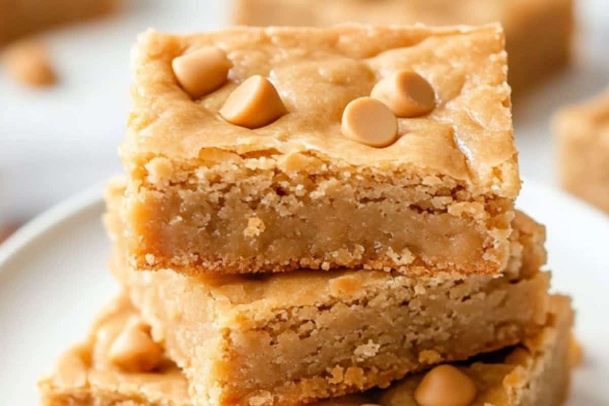 Two stacked blondies topped with delightful butterscotch chips sit on a white plate, inviting you to explore more delicious recipes. With more blondies in the background, this is a treat you must try today.