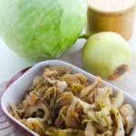 A white and red dish filled with braised cabbage sits in front of a whole green cabbage, a yellow onion, and a wooden salt container.