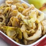 Braised cabbage with caraway seeds is served in a red and white ceramic dish, with an onion in the background.